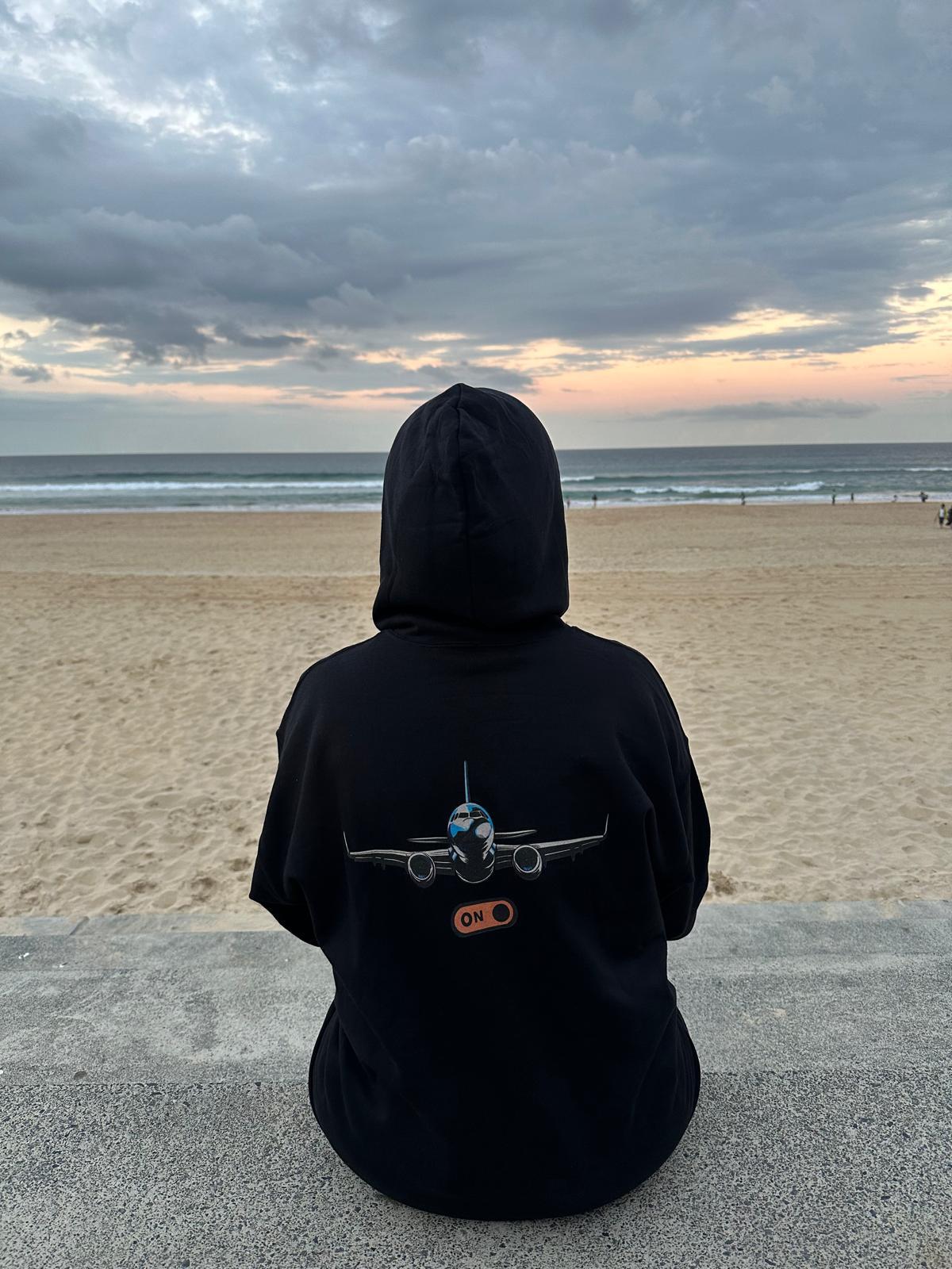 Person wearing a black hoodie with a aeroplane design sitting on a ledge overlooking a beach.