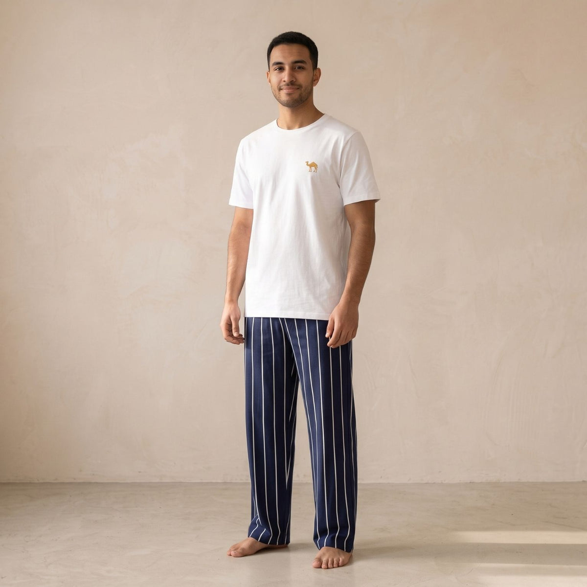 Man wearing a white T-shirt with a small camel motif and navy pants with white vertical stripes, shown from the front.