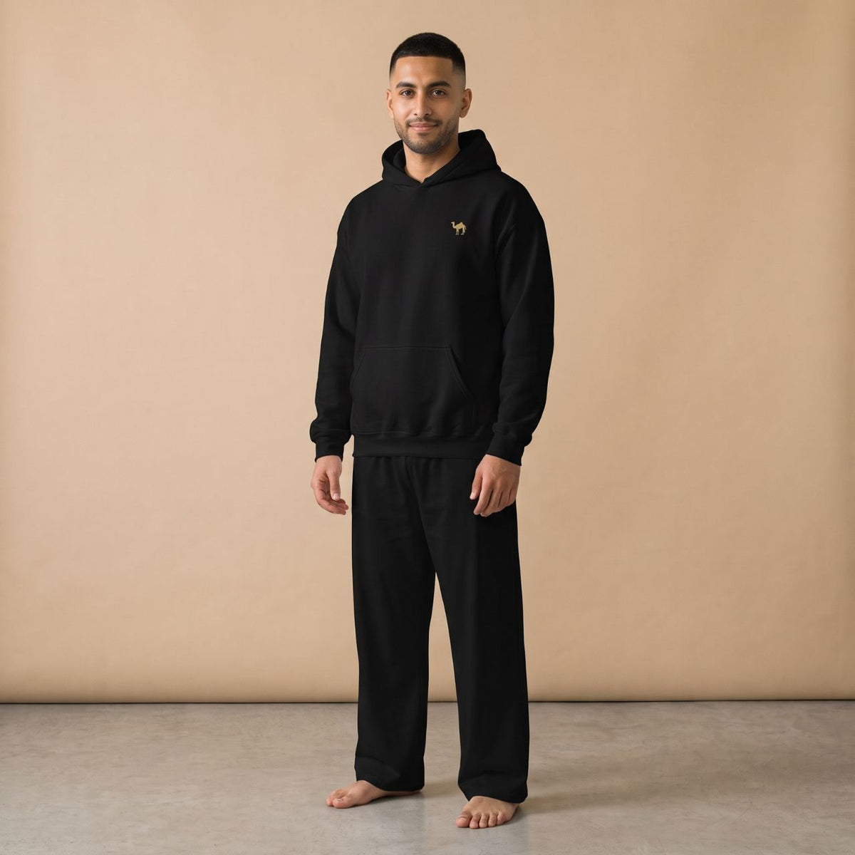 Model wearing a black hoodie with a small golden camel motif and plain black pants, front view.