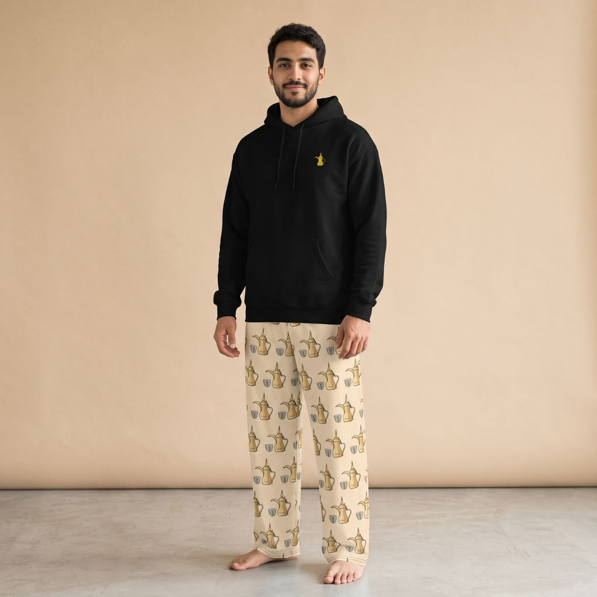 Model wearing a black hoodie with a golden dallah motif and cream pants with a dallah pot and finjan cup prints, front view.