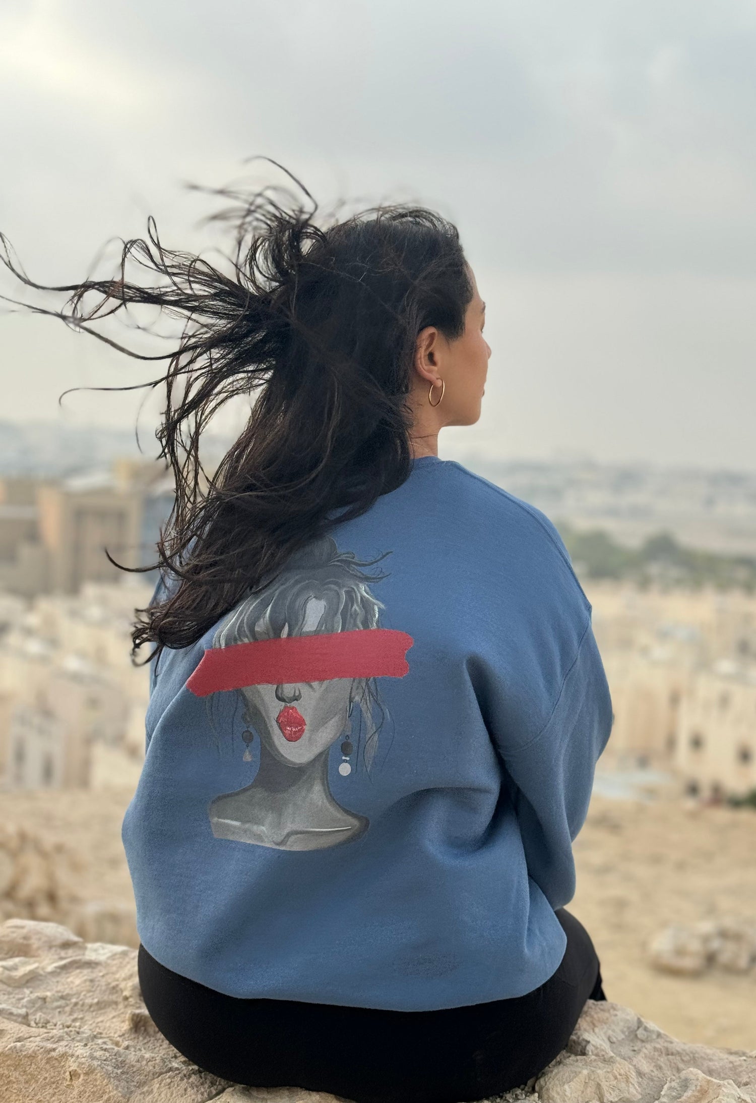 Person wearing a blue sweatshirt with a graphic design, sitting on a stone wall overlooking a scenic landscape.