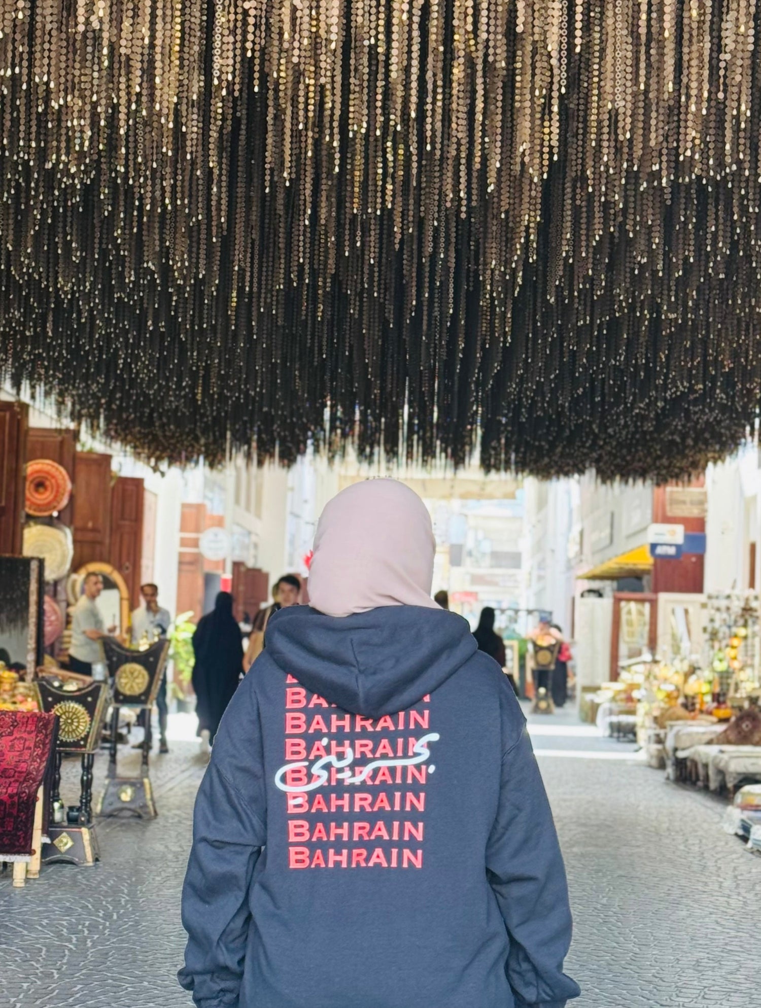 Person wearing a black hoodie with 'Bahrain' printed on it, standing under a large hanging installation.