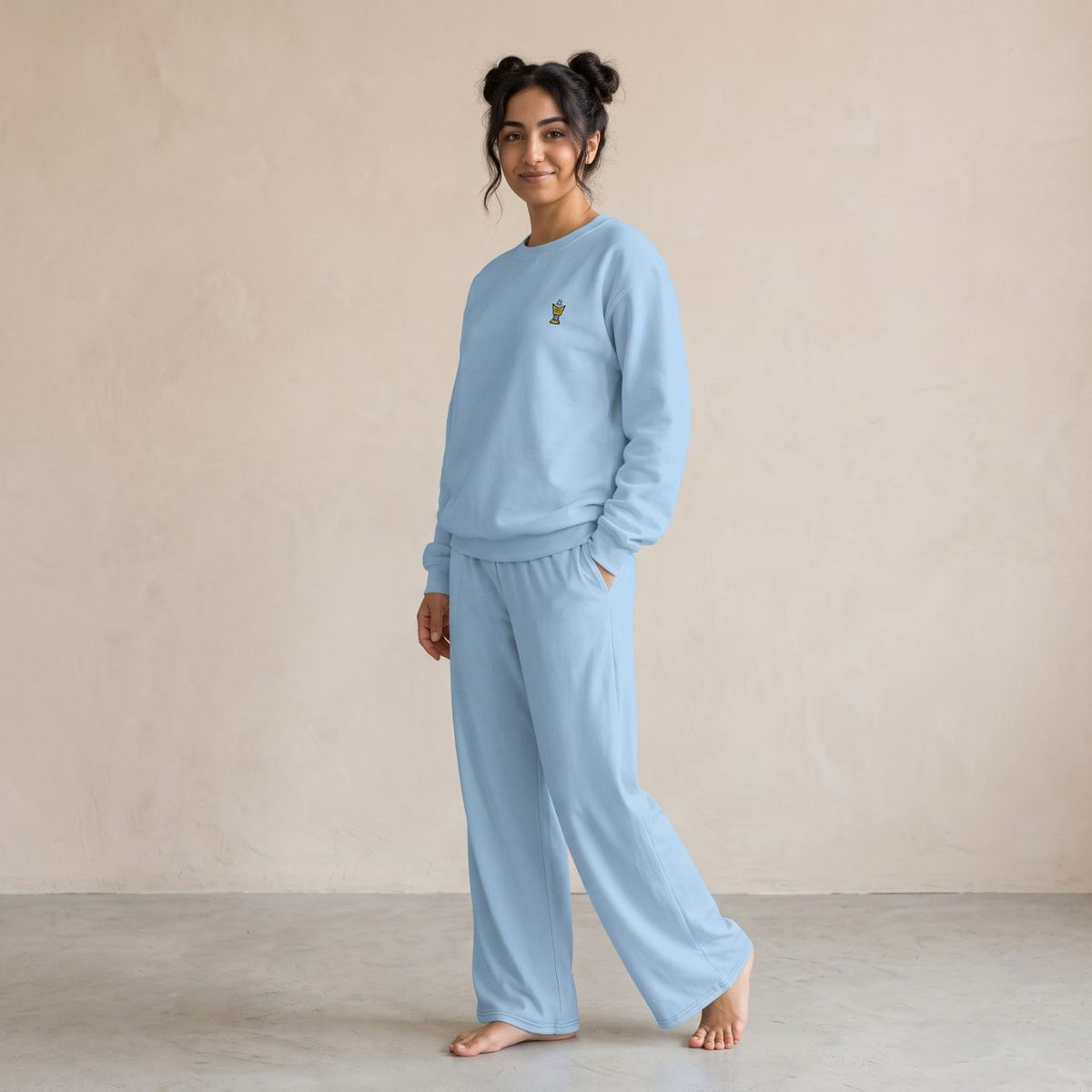 Model wearing a light blue sweatshirt with a small gold Mubkhar chest motif and matching wide-leg pants.