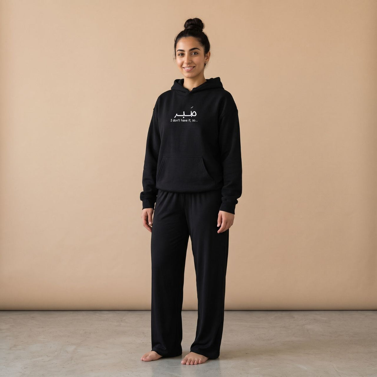 Model wearing a black hoodie with “صبر” and “I don’t have it, so…” text and plain black wide-leg pants, front view.