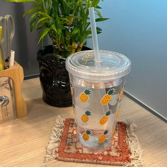 Clear tumbler with pineapple design on a coaster, surrounded by office supplies and a plant.