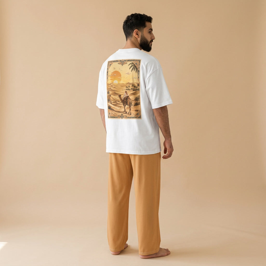 Male model viewed from behind wearing a white oversized T-shirt with a desert sunset and camel rider back print, paired with light brown wide-leg pants.