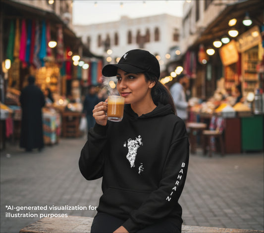 Woman in a black hoodie with a bahraini map design and cap with an embroidered bahraini map as she's holding a cup of coffee in an outdoor market setting