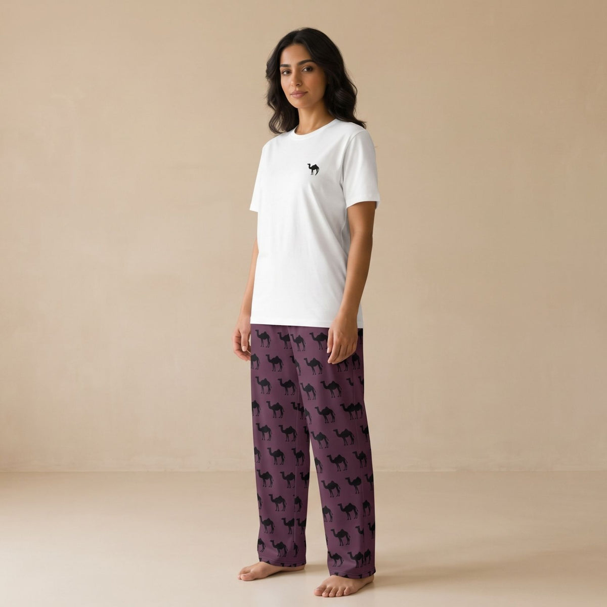 A woman stands barefoot in a studio wearing a white short-sleeve T-shirt with a small black camel print on the chest and purple pajama pants covered in a black camel print.