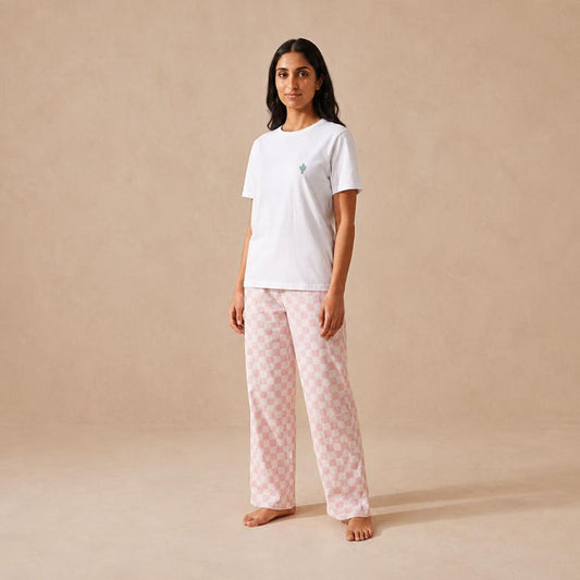 A woman stands barefoot against a warm beige studio backdrop, wearing a white short-sleeve T-shirt with a small green cactus and light pink patterned pajama pants.