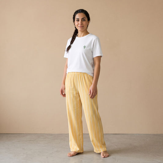 A woman stands barefoot in a studio wearing loose yellow-and-white striped pajama pants and a white T-shirt with a small cactus print on the left chest, against a plain beige wall and concrete floor.