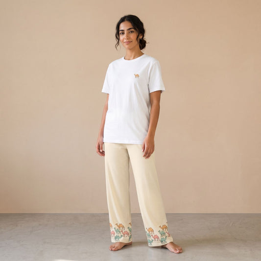 Model wearing a white T-shirt with a small camel motif and cream pants with colourful camel prints around the hems, front view.