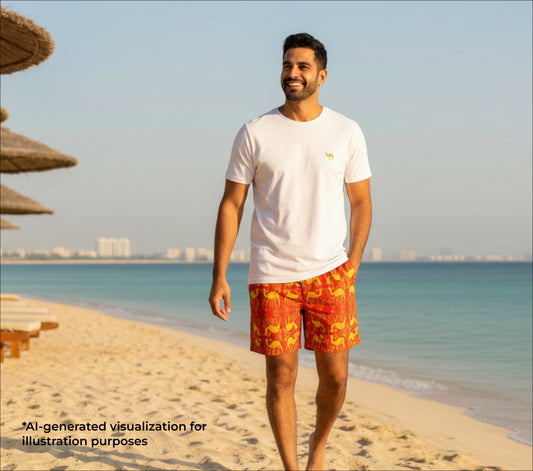 Man wearing a white t-shirt and red shorts  with an arabesque camel pattern standing on a beach with a city skyline in the background.