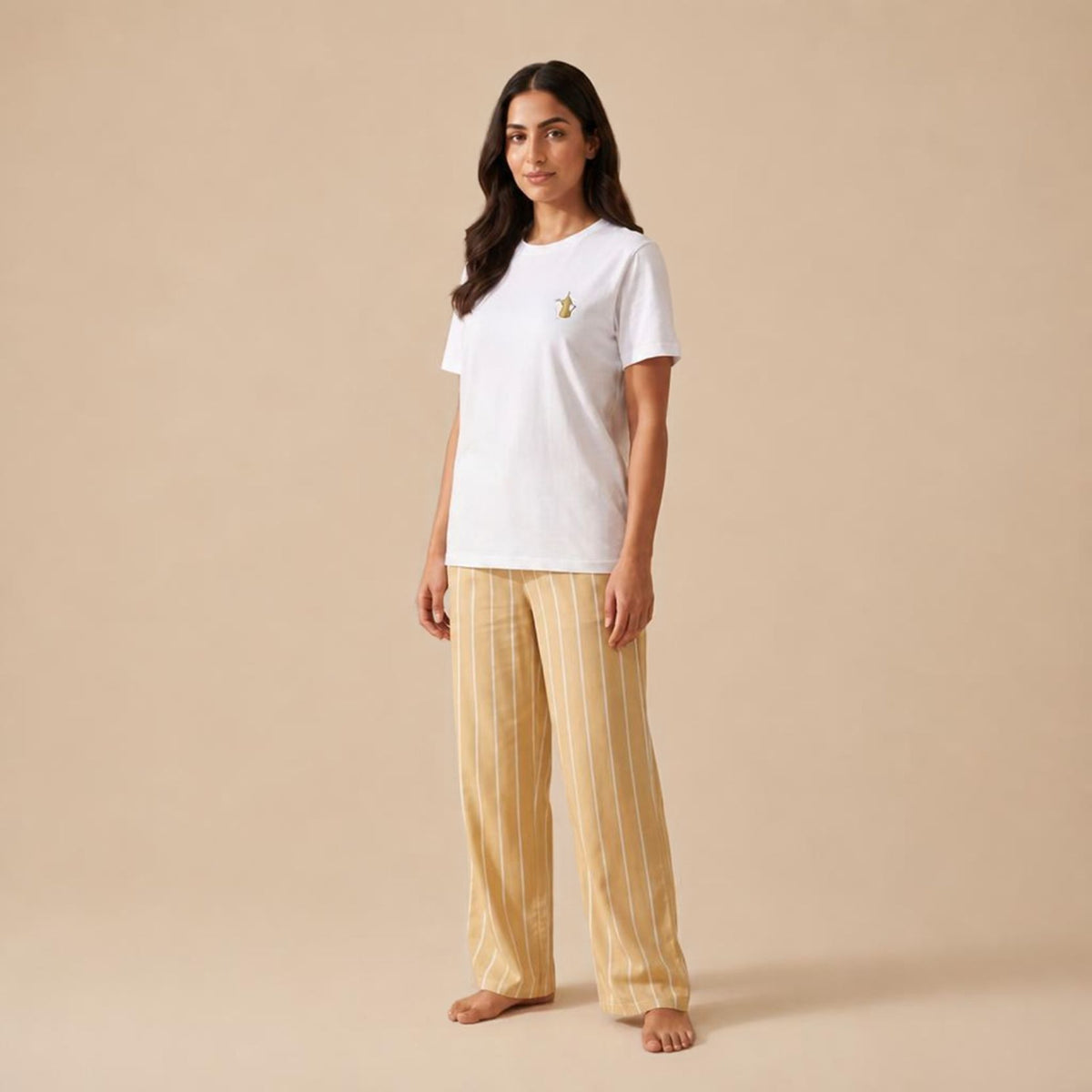 A woman stands barefoot against a plain beige studio background, wearing a white short-sleeve T-shirt with a small yellow dallat gahwa print on the left chest and light beige pinstriped pants.