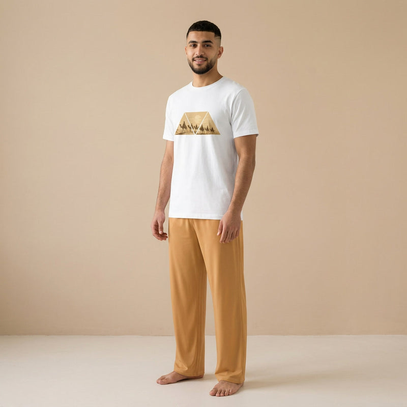 Male model wearing a white T-shirt with a golden desert caravan graphic and light brown relaxed-fit pants, shown from the front.