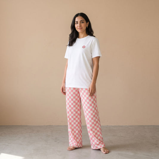 A woman stands barefoot in a studio setting, wearing a white short-sleeve t-shirt with a small pink donut print on the left chest and pink-and-white checkered wide-leg pants.