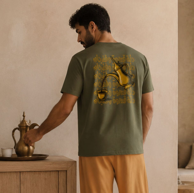 A man wearing an olive green t-shirt with a yellow coffee cup graphic on the back, paired with mustard yellow pants, standing in a warm-toned room next to a wooden table with a brass coffee pot.