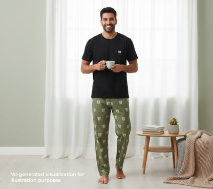 Man wearing a black t-shirt and green pants holding a cup in a room with white curtains and a small table.