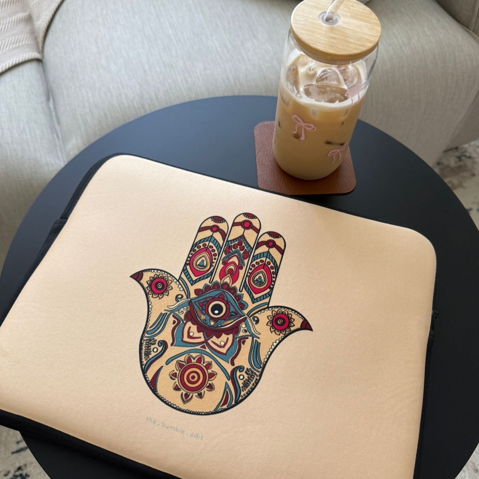 Beige laptop sleeve with Hamsa hand design on a black table, next to a glass of iced coffee.