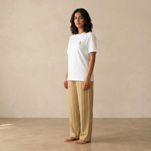 A woman stands barefoot in a studio wearing a white short-sleeve crewneck t-shirt with a small mubkhar print on the left chest, paired with beige and white vertically striped wide-leg pants.