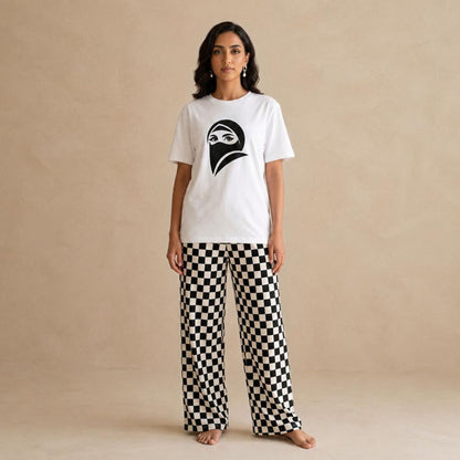 A woman stands barefoot against a warm beige textured wall, wearing a white short-sleeve T-shirt with a woman wearing a black burqa graphic on the chest and black-and-white checkered wide-leg pants.