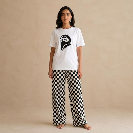 A woman stands barefoot against a warm beige textured wall, wearing a white short-sleeve T-shirt with a woman wearing a black burqa graphic on the chest and black-and-white checkered wide-leg pants.