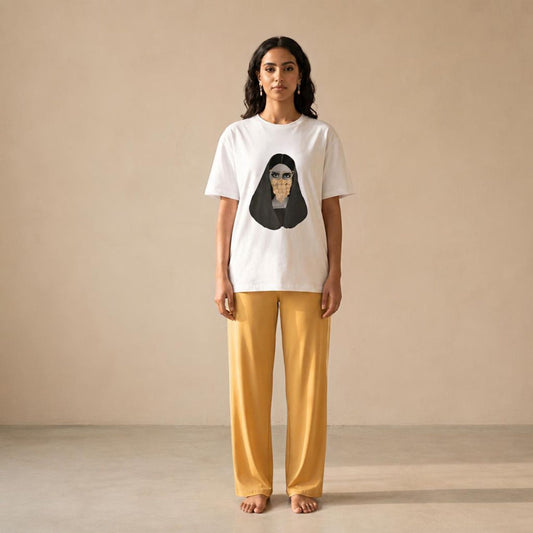 A woman stands barefoot against a plain beige studio wall wearing a white short-sleeve T-shirt with a large black portrait graphic on the front and mustard yellow wide-leg pants.